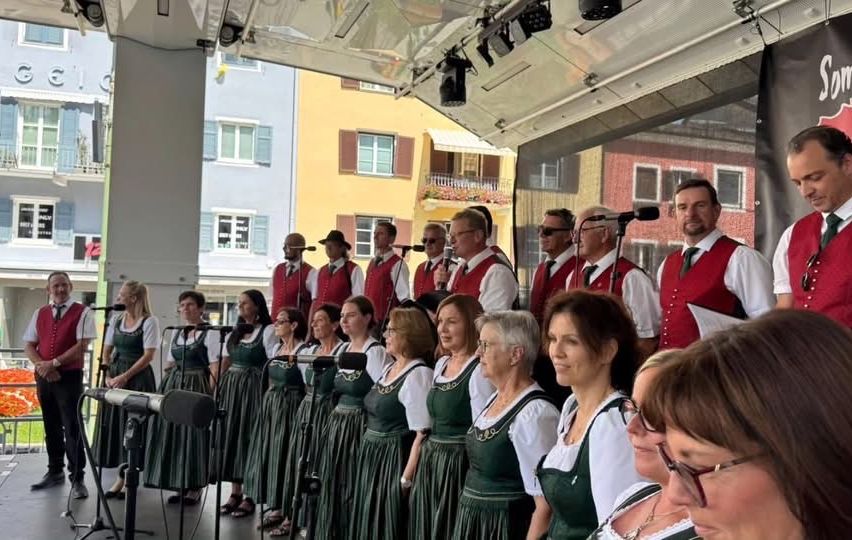Ein Chor von Menschen in traditioneller grüner und weißer Kleidung steht auf der Bühne und singt in Mikrofone. Hinter ihnen sind mehrere Gebäude zu sehen, und ein Mann steht rechts.