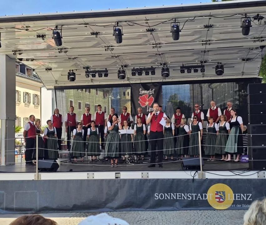 Ein Chor in traditioneller Kleidung tritt auf einer Bühne auf, mit Gebäuden im Hintergrund und einem Banner, das 'Sommerfest Lienz' besagt.