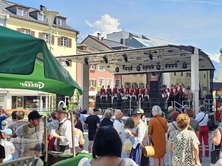 Eine Gruppe von Menschen in traditioneller Kleidung tritt auf einer Bühne vor einer Menge in einem Stadtplatz auf.