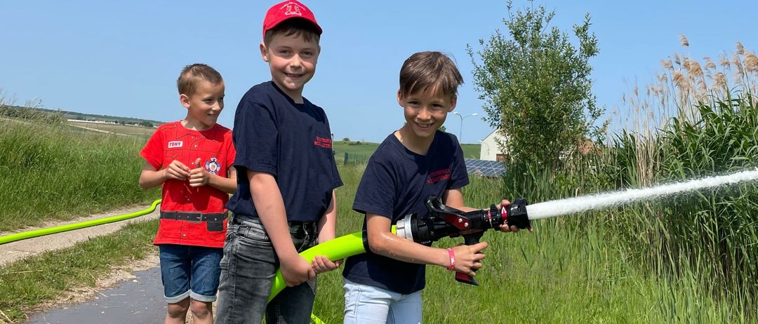 Drei Jungen spielen mit einem Schlauch auf einer Wiese. Ein Junge sprüht Wasser, während die anderen beiden zusehen. Sie stehen auf einer Wiese mit hohem Gras und einem Haus in der Ferne.
