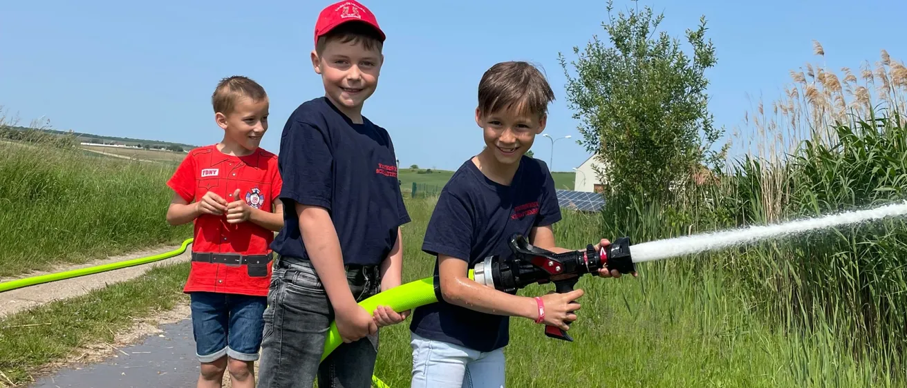 Drei Jungen spielen mit einem Schlauch auf einer Wiese. Ein Junge sprüht Wasser, während die anderen beiden zusehen. Sie stehen auf einer Wiese mit hohem Gras und einem Haus in der Ferne.
