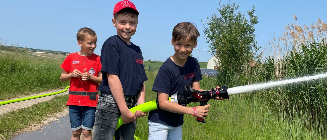 Drei Jungen spielen mit einem Schlauch auf einer Wiese. Ein Junge sprüht Wasser, während die anderen beiden zusehen. Sie stehen auf einer Wiese mit hohem Gras und einem Haus in der Ferne.