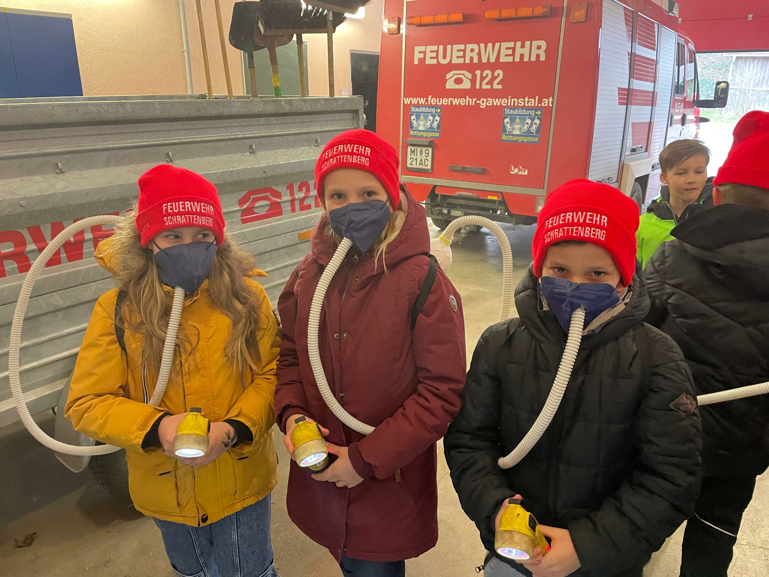 Drei Kinder, jedes mit einem roten Hut und einer Gesichtsmaske, stehen vor einem Feuerwehrauto und halten Taschenlampen in der Hand. Das Feuerwehrauto hat ein rotes und weißes Farbschema.