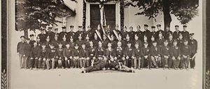 Ein Schwarz-Weiß-Foto einer Gruppe von Feuerwehrleuten in Uniform, die vor einem Gebäude mit Flaggen und Bäumen im Hintergrund stehen.