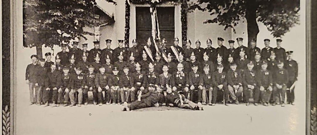 Ein Schwarz-Weiß-Foto einer Gruppe von Feuerwehrleuten in Uniform, die vor einem Gebäude mit Flaggen und Bäumen im Hintergrund stehen.