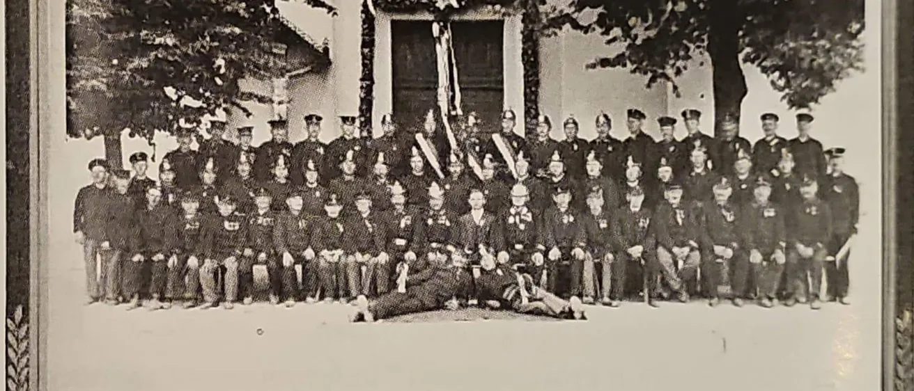 Ein Schwarz-Weiß-Foto einer Gruppe von Feuerwehrleuten in Uniform, die vor einem Gebäude mit Flaggen und Bäumen im Hintergrund stehen.