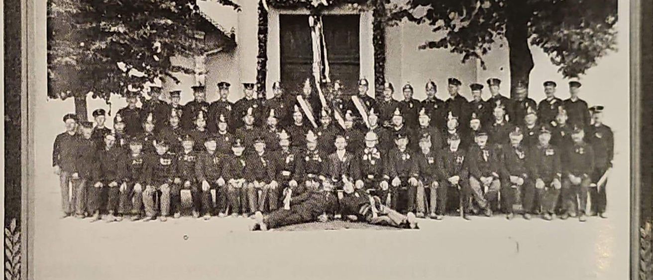 Ein Schwarz-Weiß-Foto einer Gruppe von Feuerwehrleuten in Uniform, die vor einem Gebäude mit Flaggen und Bäumen im Hintergrund stehen.