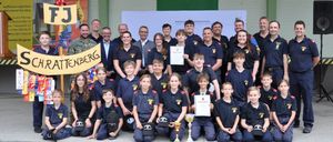 Eine Gruppe junger Menschen in blauen Uniformen mit einigen Erwachsenen posiert für ein Foto mit Trophäen und Urkunden. Sie stehen auf einer Betonfläche.