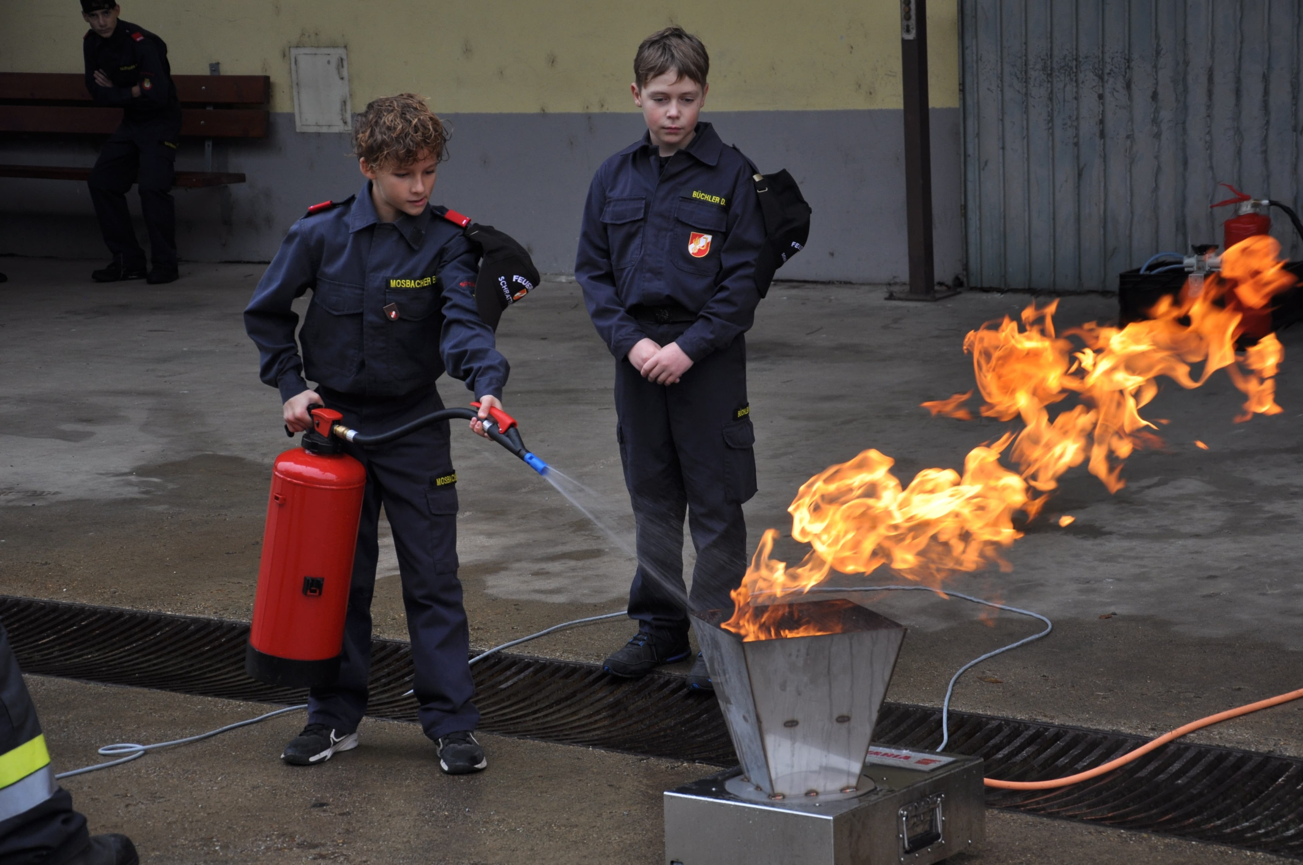 Zwei Jungen in Uniformen verwenden einen Feuerlöscher, um ein Feuer in einem Metallbehälter zu löschen.