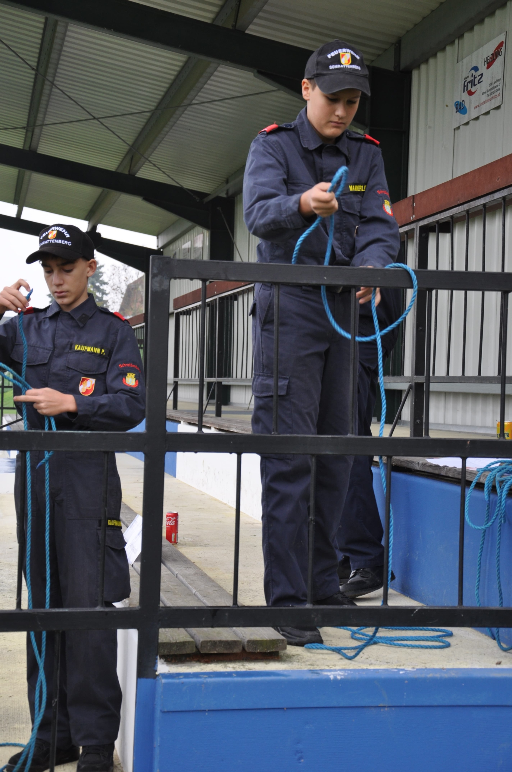 Zwei junge Männer in marineblauen Uniformen binden blaue Seile um ein Metalltor, möglicherweise bei einer Trainingsveranstaltung.