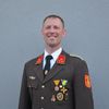 Ein Mann in Uniform steht gegen eine graue Wand und lächelt. Er trägt eine Krawatte und Medaillen, die seinen Dienst zeigen.