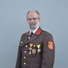 Ein Mann in Uniform steht vor einer grauen Wand. Er hat eine Pfeife im Mund und trägt eine Brille. Er hat Medaillen auf der linken Seite.