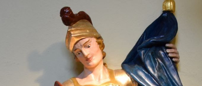 Eine Statue einer Frau in goldenen und roten Gewändern, die einen Becher hält, mit einem blauen Umhang um ihre Schultern. Sie steht an einer hellen Wand.