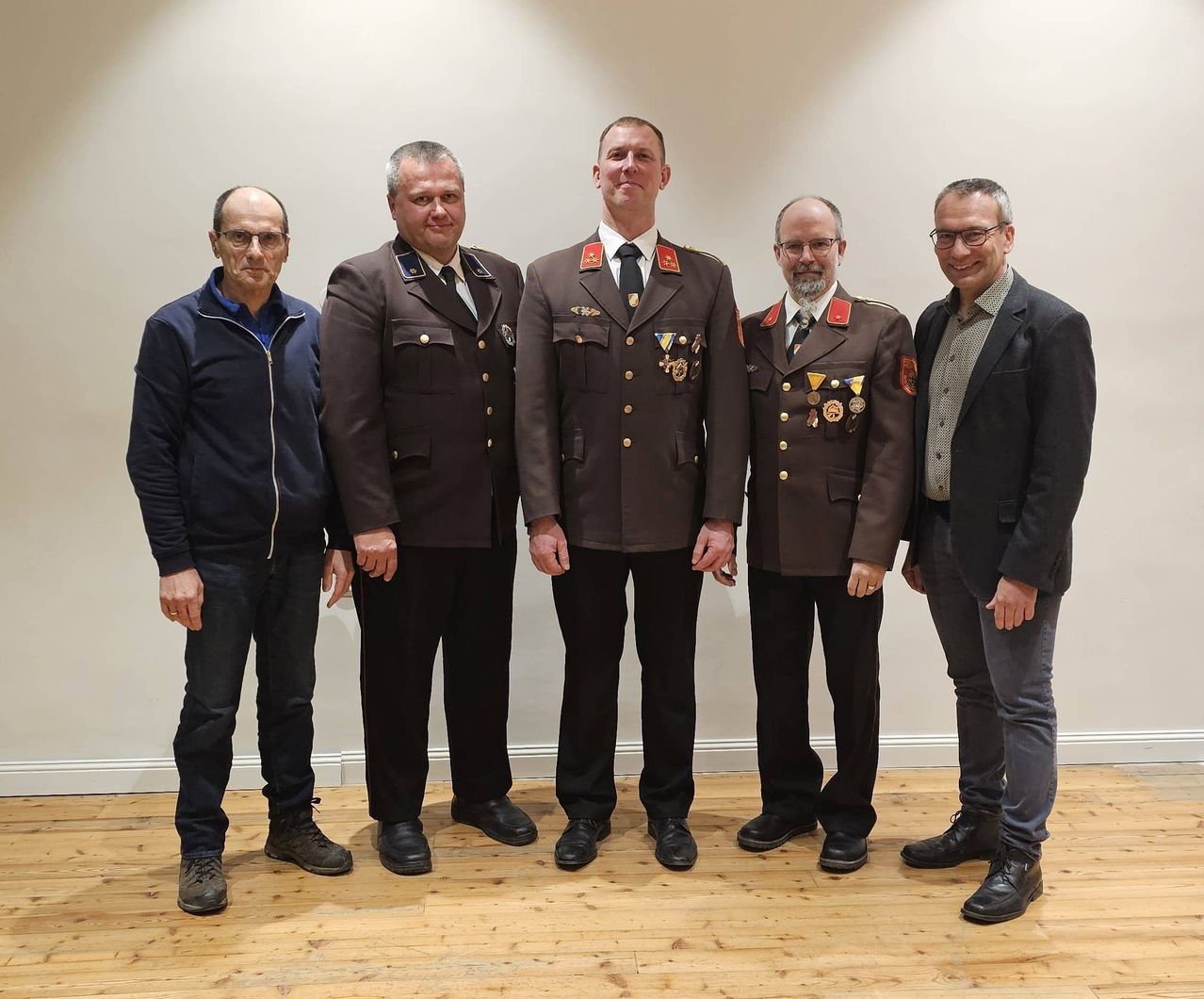 Fünf Männer stehen zusammen, wahrscheinlich für ein Foto, in formeller Kleidung. Drei tragen Militäruniformen, einer trägt einen Anzug und einer trägt eine Jacke. Sie lächeln und stehen auf einem Holzboden.