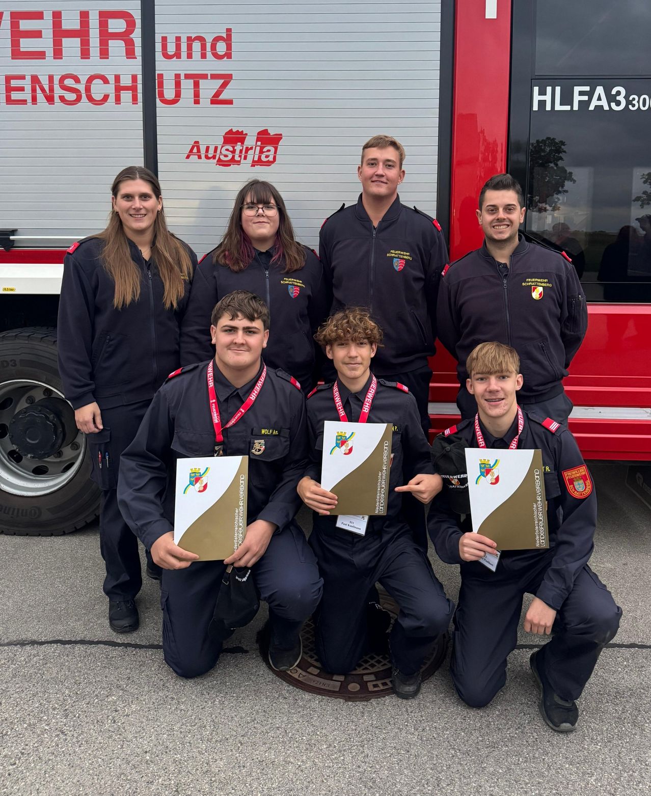 Sieben Personen in Feuerwehruniformen posieren vor einem Feuerwehrauto mit der Aufschrift Austria und HLFA330. Sie halten Urkunden in den Händen.