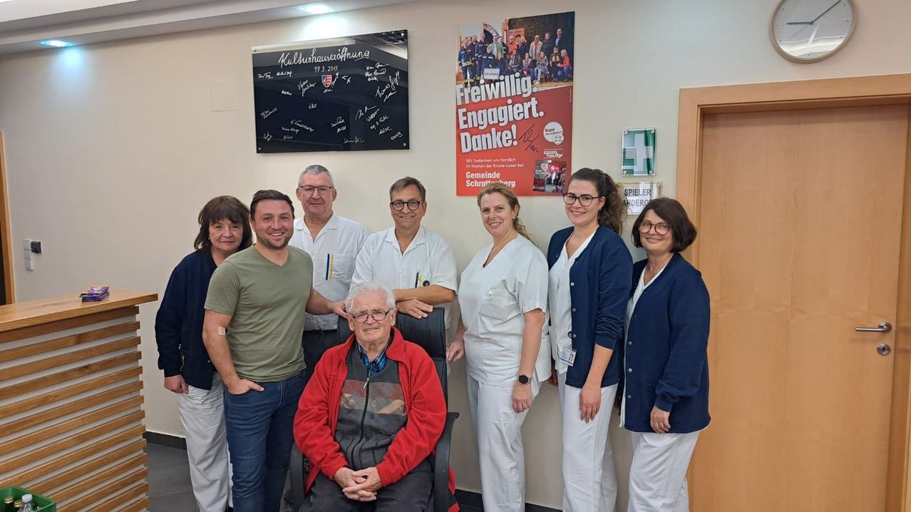 Eine Gruppe von Menschen steht in einem Raum mit einem Mann im Stuhl, der lächelt. Dahinter befindet sich ein Schild mit der Aufschrift 'Freiwillig. Engagirt. Danke!' mit einem Logo und Text. Ein weiteres Schild ist an der Wand befestigt.