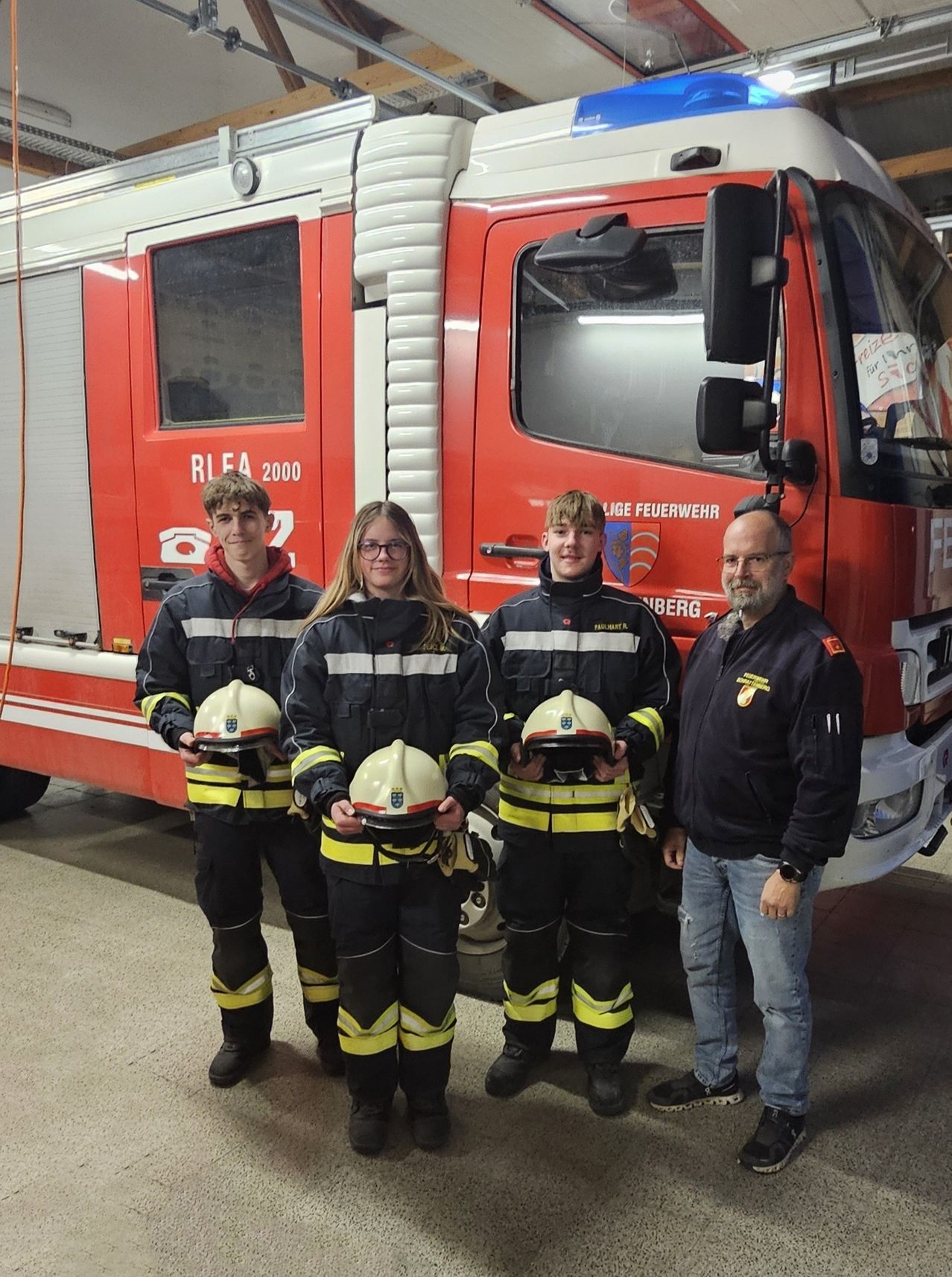 Drei Feuerwehrleute und ein älterer Mann stehen vor einem Feuerwehrauto. Die Feuerwehrleute tragen Uniformen und halten Helme. Der Mann trägt eine Brille und eine Uhr.