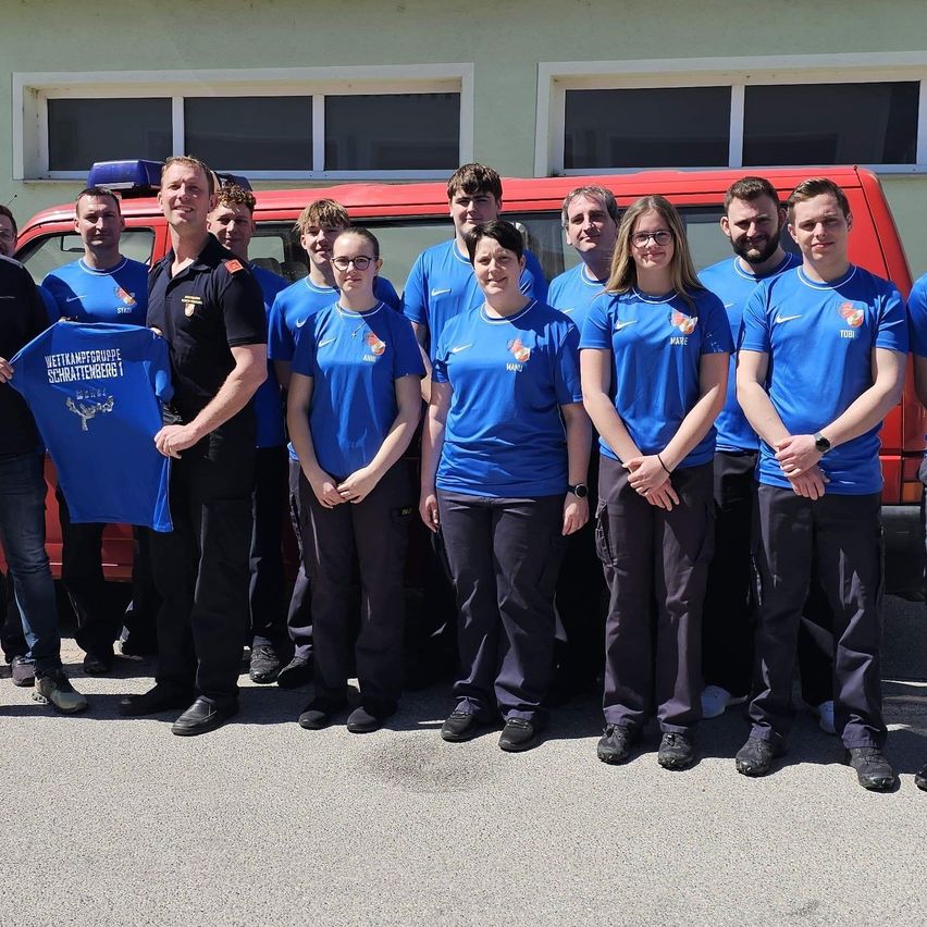 Eine Gruppe von Menschen in blauen T-Shirts und schwarzen Hosen steht vor einem roten Van, wobei eine Person ein blaues T-Shirt mit einem Logo hält.