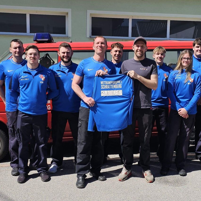 Eine Gruppe von Feuerwehrleuten posiert mit einem blauen Shirt. Ein Mann hält ein blaues Shirt mit Text. Sie stehen vor einem roten Fahrzeug.