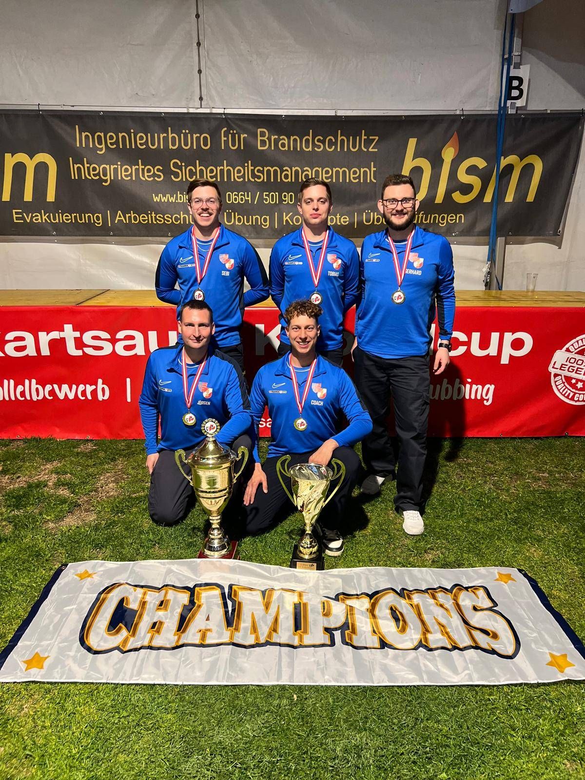 Fünf Männer in blauen Uniformen posieren mit ihren Medaillen und Pokalen auf einem Rasenfeld. Sie stehen vor einem Banner mit der Aufschrift 'Champions'. Dahinter befindet sich eine Bühne mit einem Banner.