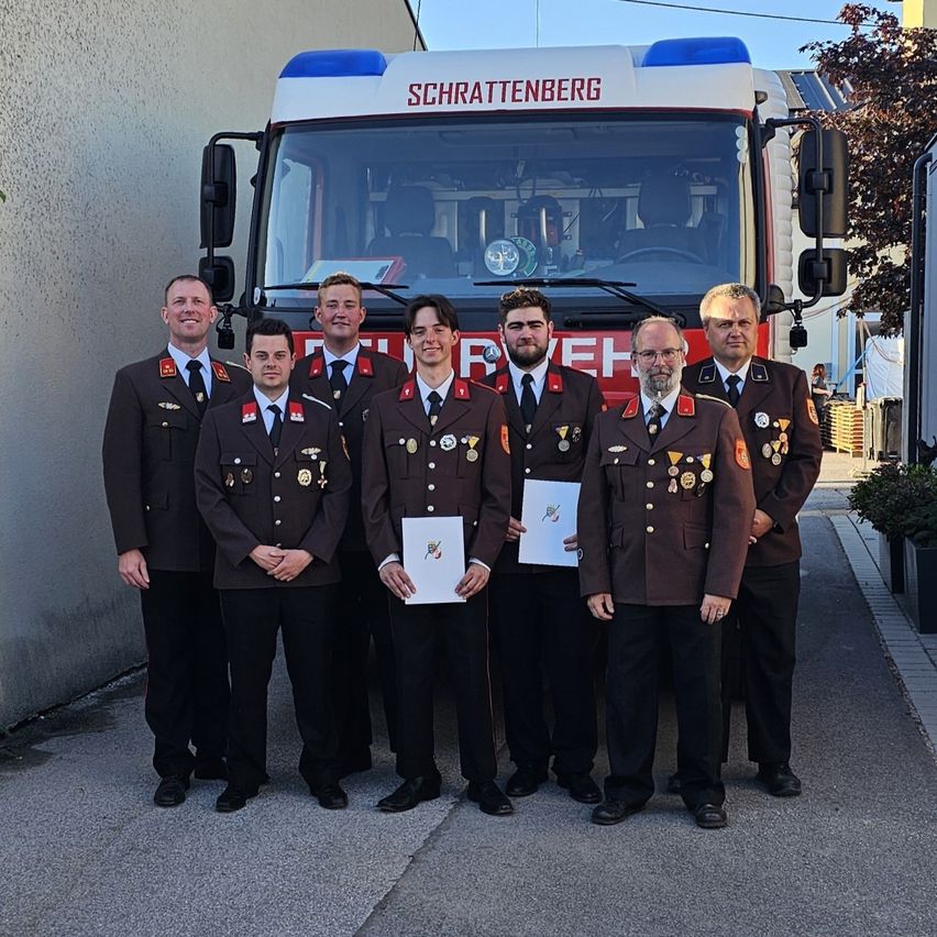 Sieben Feuerwehrleute stehen vor einem Feuerwehrwagen namens Schrattenberg, halten Zertifikate in der Hand und tragen Uniformen.