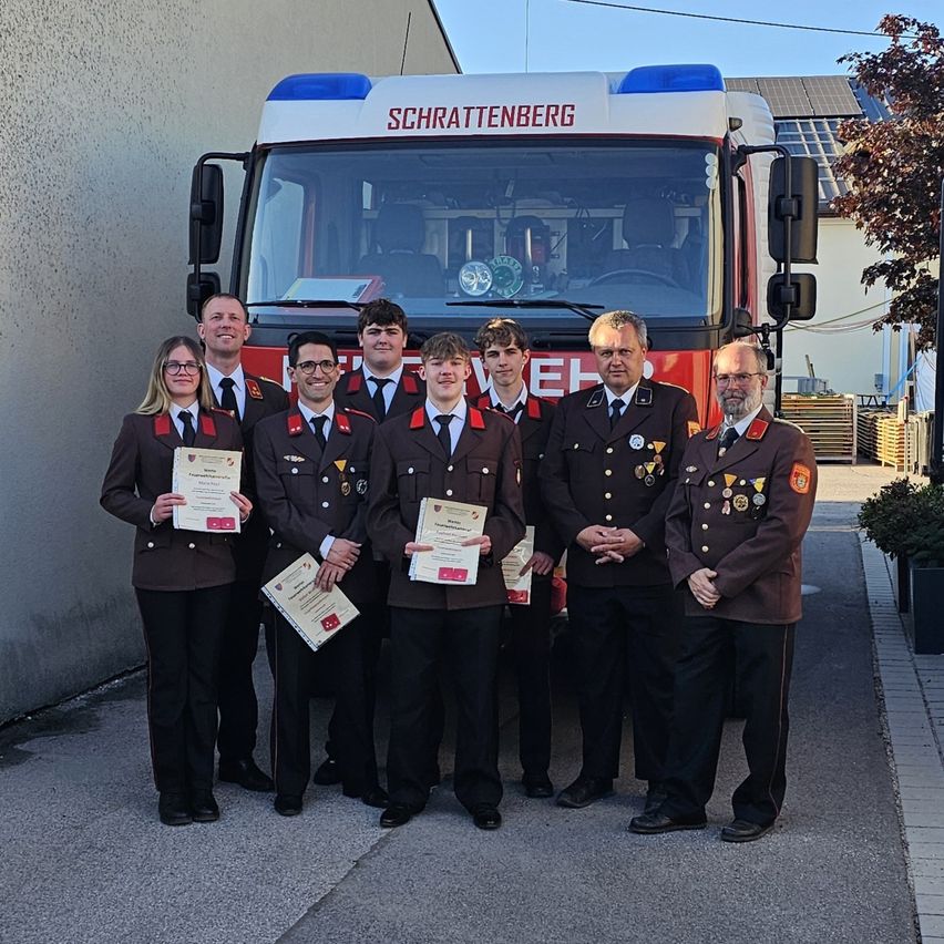 Eine Gruppe von Menschen in Uniformen steht vor einem Feuerwehrauto und hält Zertifikate in den Händen. Das Auto ist mit Schrattenberg beschriftet. Dahinter befindet sich ein Gebäude mit Solarpanelen.