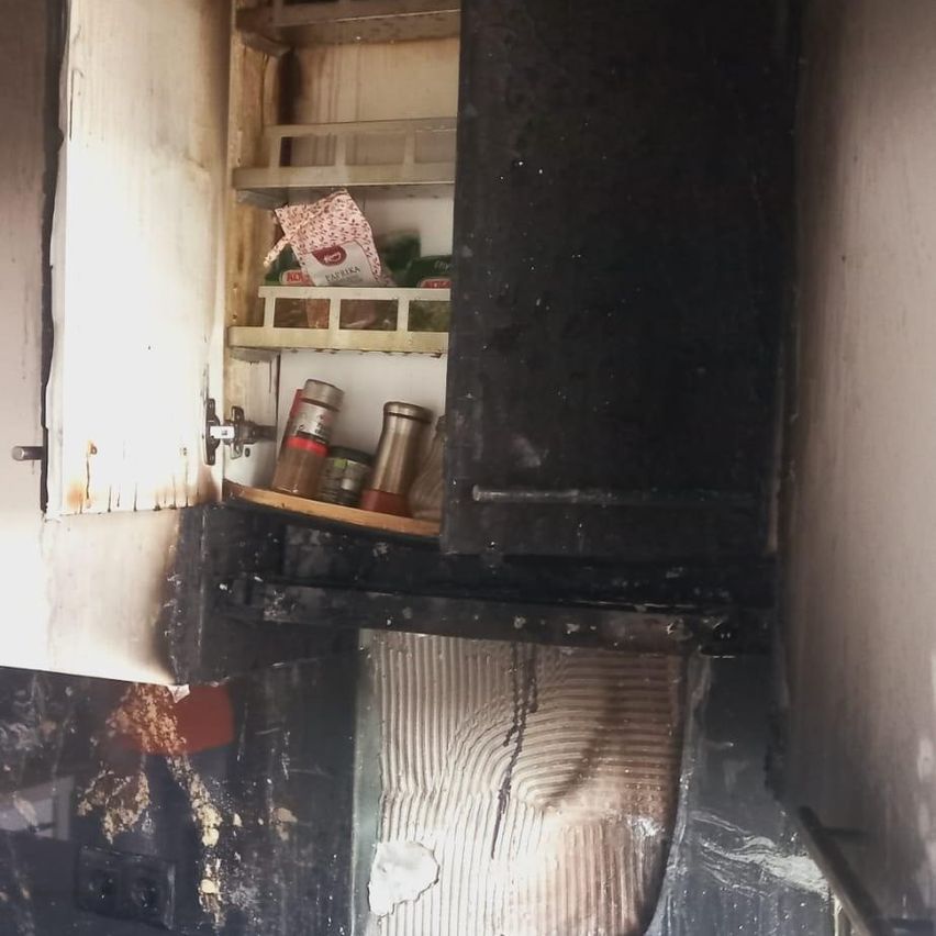 Ein alter, verbrannter Schrank mit offenen Türen zeigt einige Dosen und eine Packung im Regal. Der Schrank und die Wand sind verkohlt.