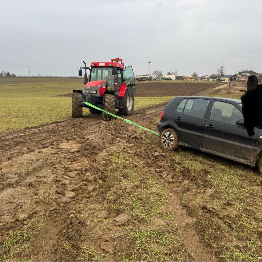 Ein roter Traktor zieht ein schwarzes Auto aus schlammigem Boden in einem Feld mit einem bewölkten Himmel darüber.