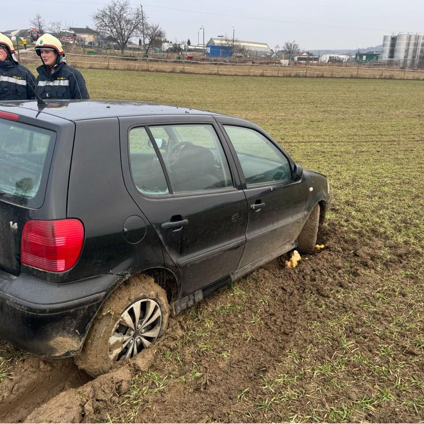 Ein schwarzes Auto ist im Schlamm stecken geblieben, zwei Feuerwehrleute stehen dahinter, einer trägt einen Helm. Das Feld ist von einem Zaun umgeben, und es gibt Bäume und Gebäude in der Ferne.