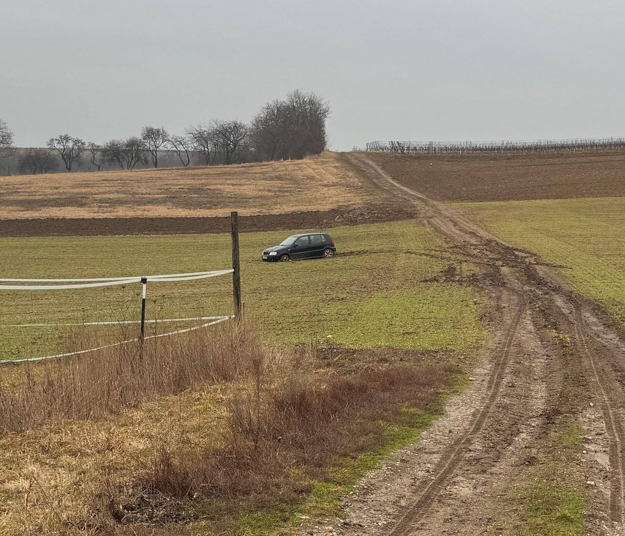 Ein schwarzes Auto steckt im schlammigen Boden in der Nähe eines Feldwegs in einem weiten, offenen Feld mit Bäumen in der Ferne.