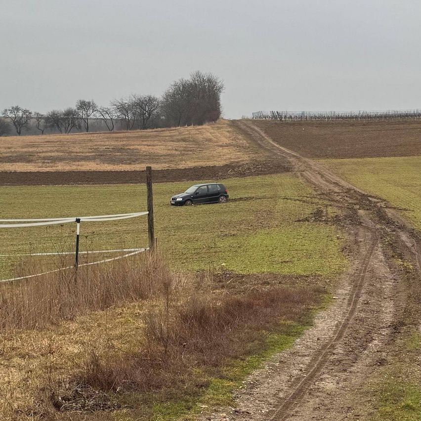 Ein schwarzes Auto steckt im schlammigen Boden in der Nähe eines Feldwegs in einem weiten, offenen Feld mit Bäumen in der Ferne.
