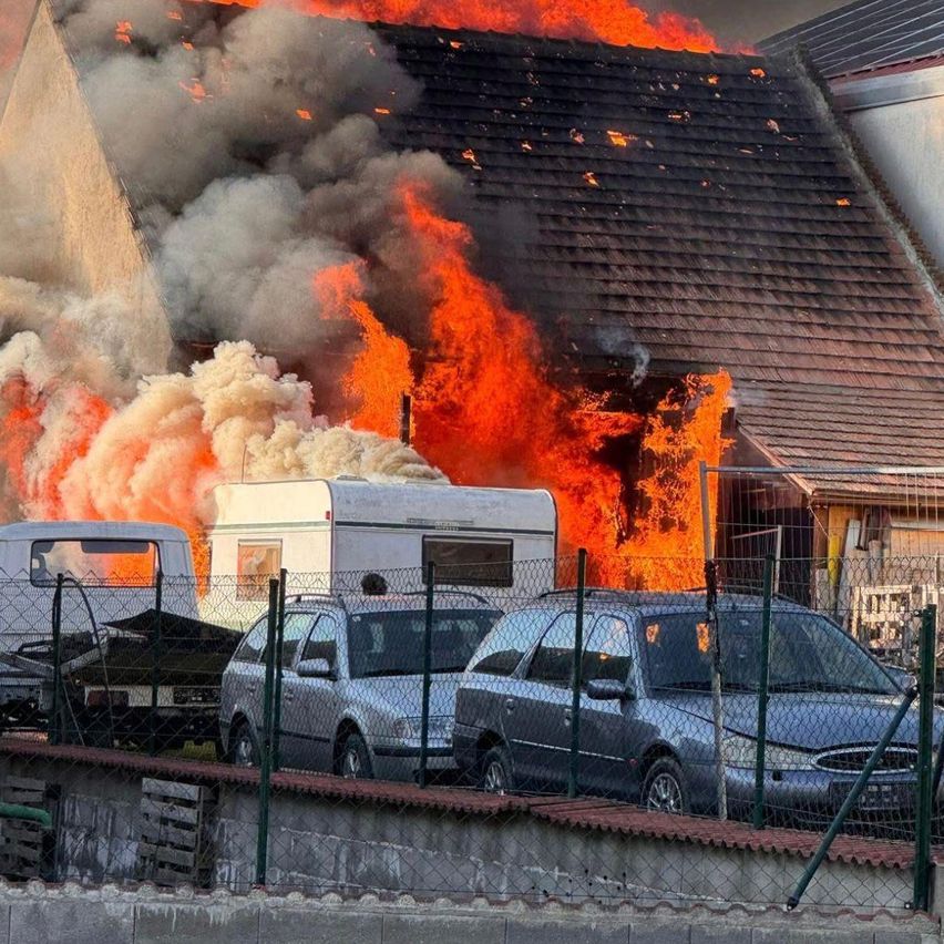 Ein Haus in Flammen, mit dickem Rauch, umgeben von geparkten Fahrzeugen, einschließlich eines Wohnwagens, und einem Zaun aus Kettengliedern.