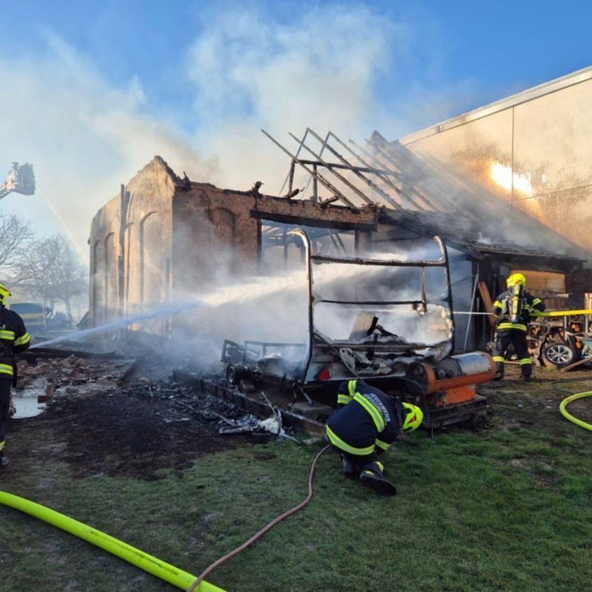 Feuerwehrleute löschen ein Feuer in einem Gebäude mit einem beschädigten Dach. Zwei Feuerwehrleute stehen in der Nähe, einer hält einen Schlauch. Ein Feuerwehrwagen befindet sich auf der linken Seite.