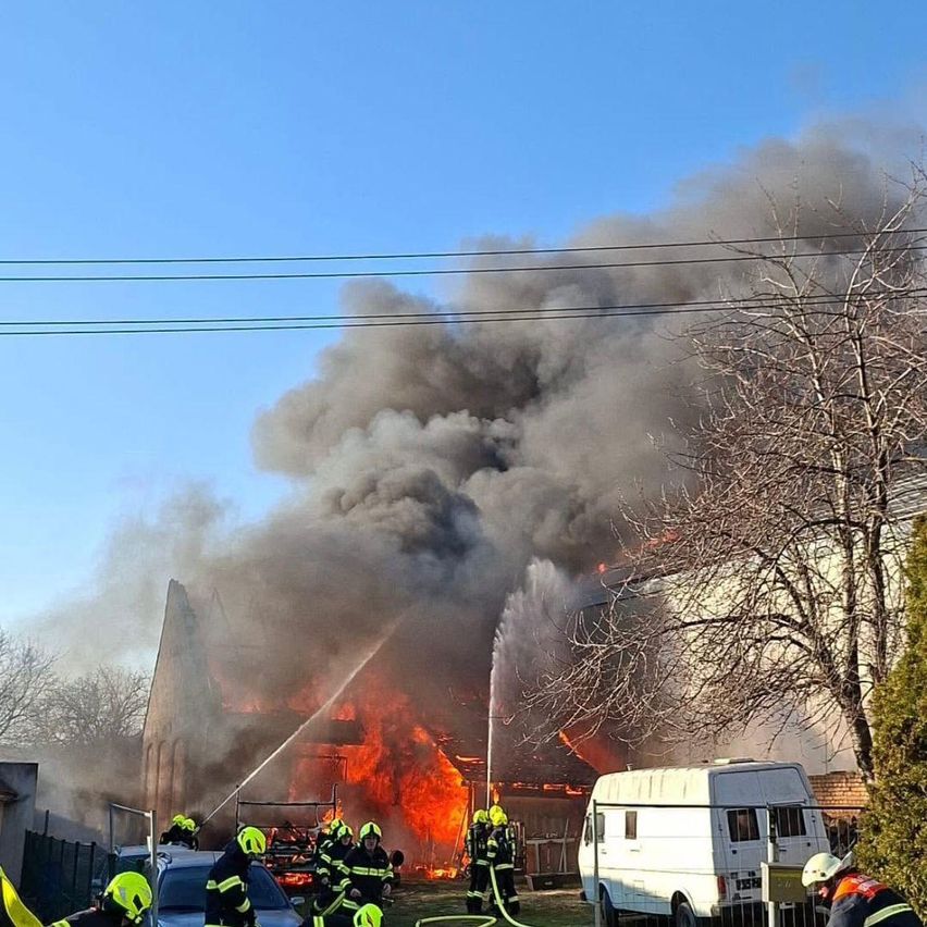 Feuerwehrleute löschen ein großes Feuer an einem Gebäude. Der Brand produziert viel Rauch. Ein weißer Van ist in der Nähe geparkt.
