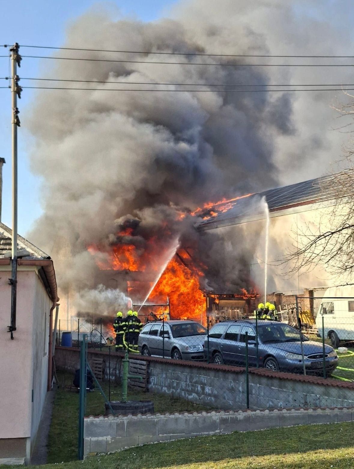 Feuerwehrleute löschen ein Feuer, das ein Gebäude verzehrt. Ein Feuerwehrwagen und zwei Autos sind in der Nähe geparkt.