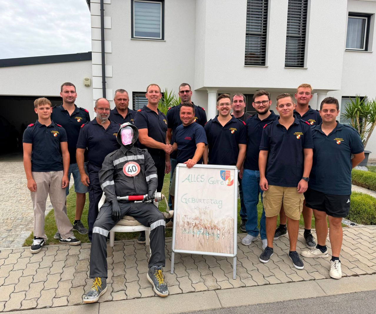 Eine Gruppe von Feuerwehrleuten in Uniform posiert für ein Foto vor einem weißen Gebäude. Eine Person sitzt in einer Feuerwehruniform.