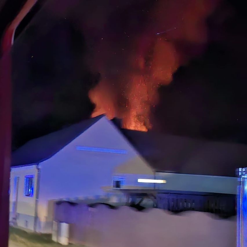 Ein Haus ist teilweise im Vordergrund sichtbar, mit einem großen Feuer im Hintergrund bei Nacht.