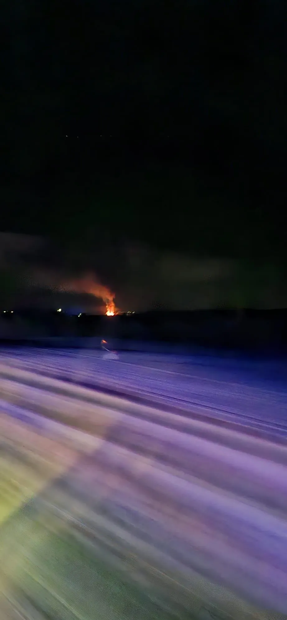 Ein nächtlicher Blick auf ein großes Feuer in der Ferne, mit dunklem Himmel und schwachen Stadtlichtern.