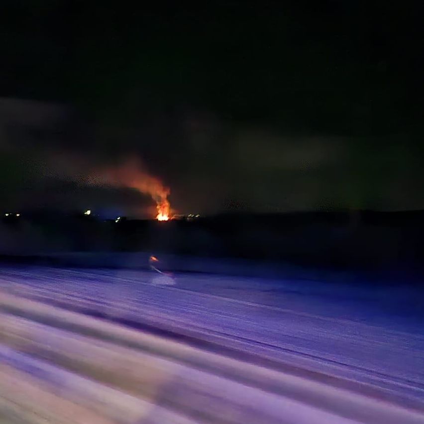 Ein nächtlicher Blick auf ein großes Feuer in der Ferne, mit dunklem Himmel und schwachen Stadtlichtern.