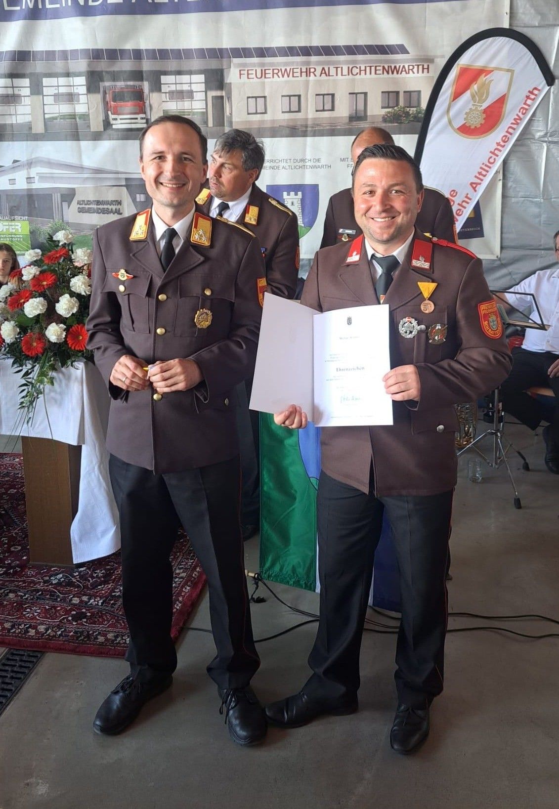 Zwei Männer in Militäruniformen, einer hält eine Urkunde in der Hand, stehen neben einer Fahne. Dahinter steht ein Banner mit der Aufschrift 'Feuerwehr Attlichtung'. Eine Frau im Hintergrund schaut zur Kamera.