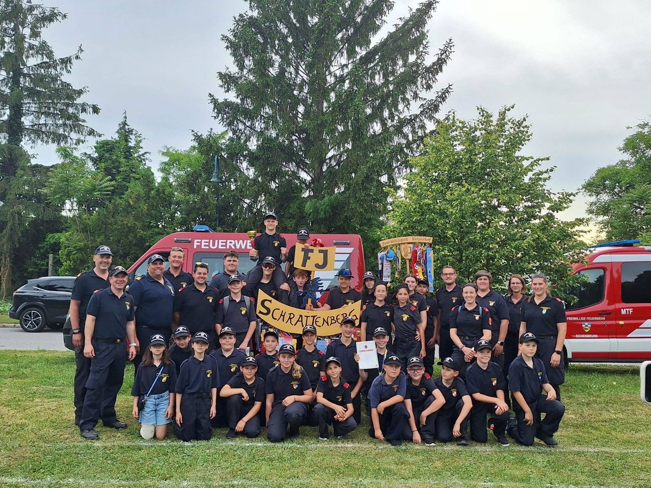 Eine Gruppenaufnahme von Feuerwehrleuten in Uniform, einige halten ein Banner mit Text, stehen auf einem Feld mit einem Feuerwehrwagen im Hintergrund.