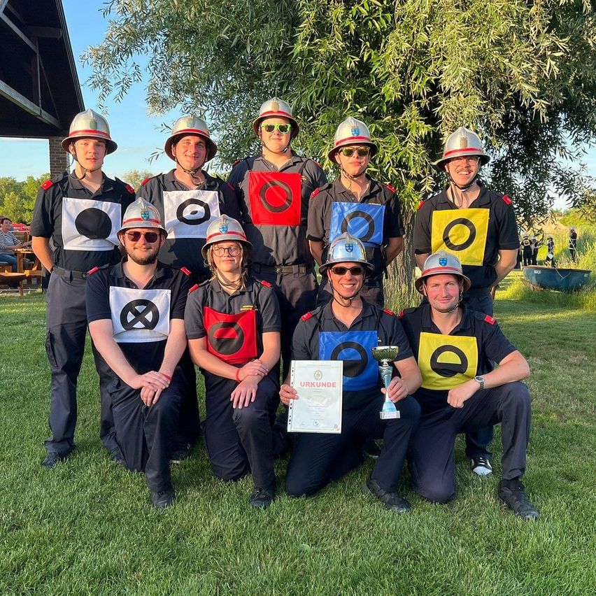 Eine Gruppe von Leuten in Uniform posiert für ein Foto. Eine Person hält ein Zertifikat. Sie tragen alle Helme und Sonnenbrillen. Der Boden ist mit Gras bedeckt.
