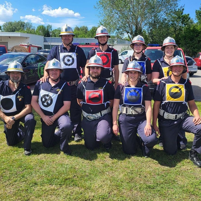 Eine Gruppe Feuerwehrleute in Uniformen und Helmen posiert für ein Foto auf dem Rasen, mit Bäumen und geparkten Fahrzeugen im Hintergrund.