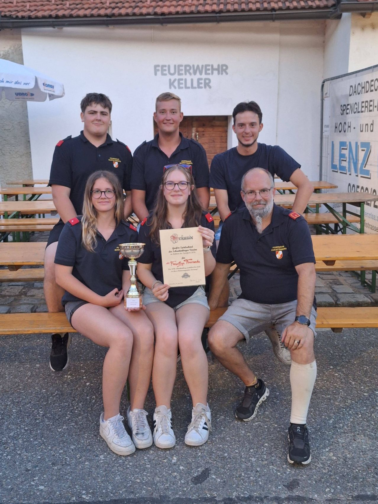 Eine Gruppe Feuerwehrleute in Uniform sitzt auf einer Bank, zwei Frauen halten einen Pokal und ein Zertifikat. Dahinter steht ein Gebäude mit dem Schriftzug 'Feuerwehr Keller'.