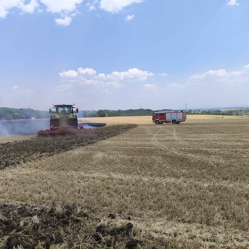 Ein Traktor pflügt ein Feld, während ein Feuerwehrwagen in der Nähe geparkt ist. Der Himmel ist klar mit einigen Wolken.