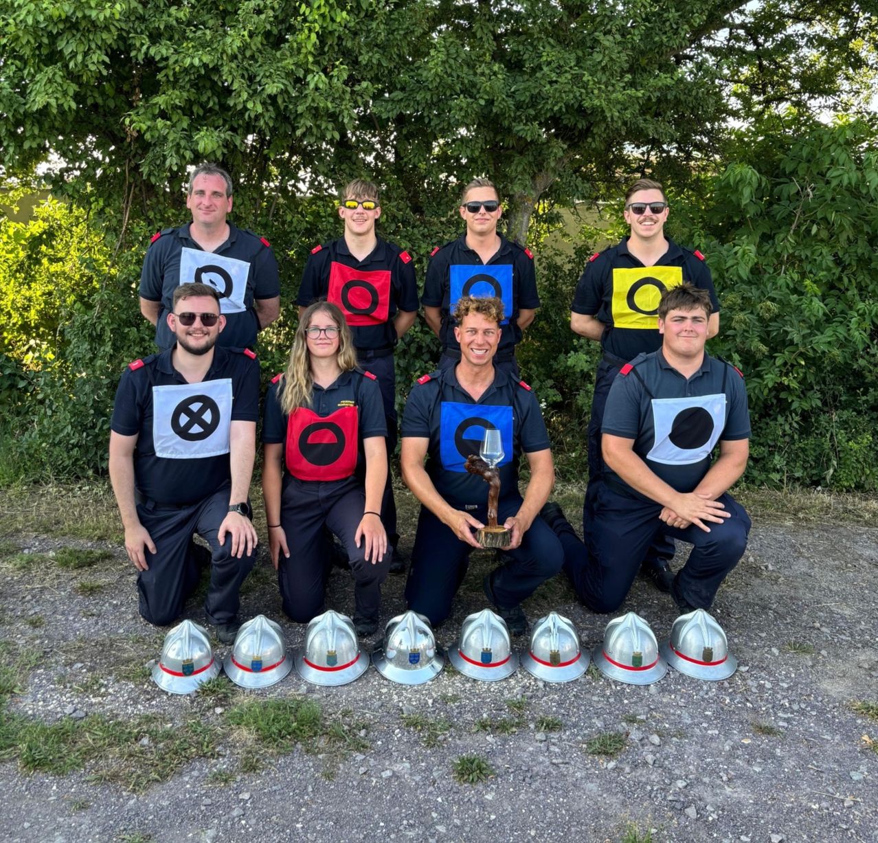 Eine Gruppe von Feuerwehrleuten, sowohl Männer als auch Frauen, posiert für ein Foto mit ihren Helmen und einer Trophäe, in Uniformen mit kreisförmigen Abzeichen.