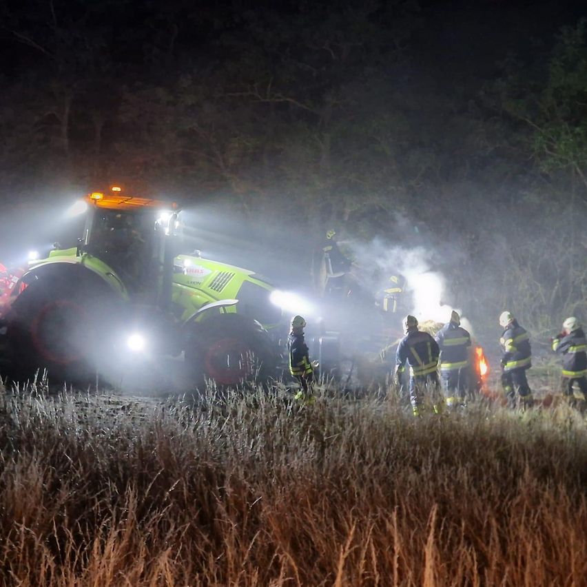 Feuerwehrleute stehen um ein brennendes Fahrzeug auf einem Feld bei Nacht, mit einem Traktor in der Nähe.