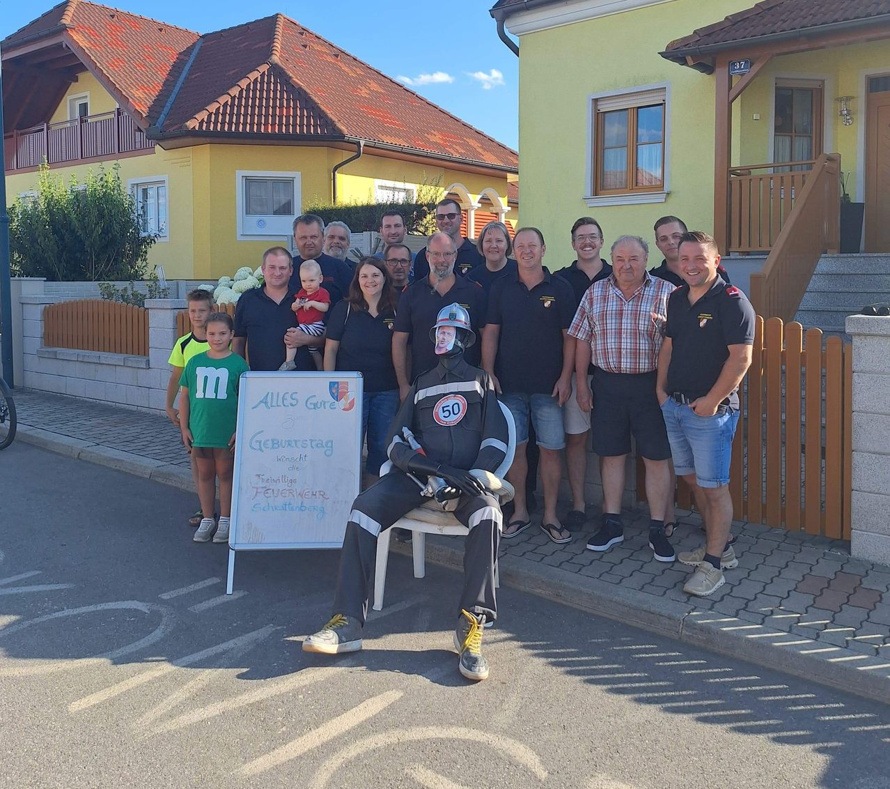 Eine Gruppe von Menschen, einschließlich Erwachsener und Kindern, posiert für ein Foto vor einem Haus. Eine Person in einer Feuerwehruniform sitzt auf einem Stuhl.