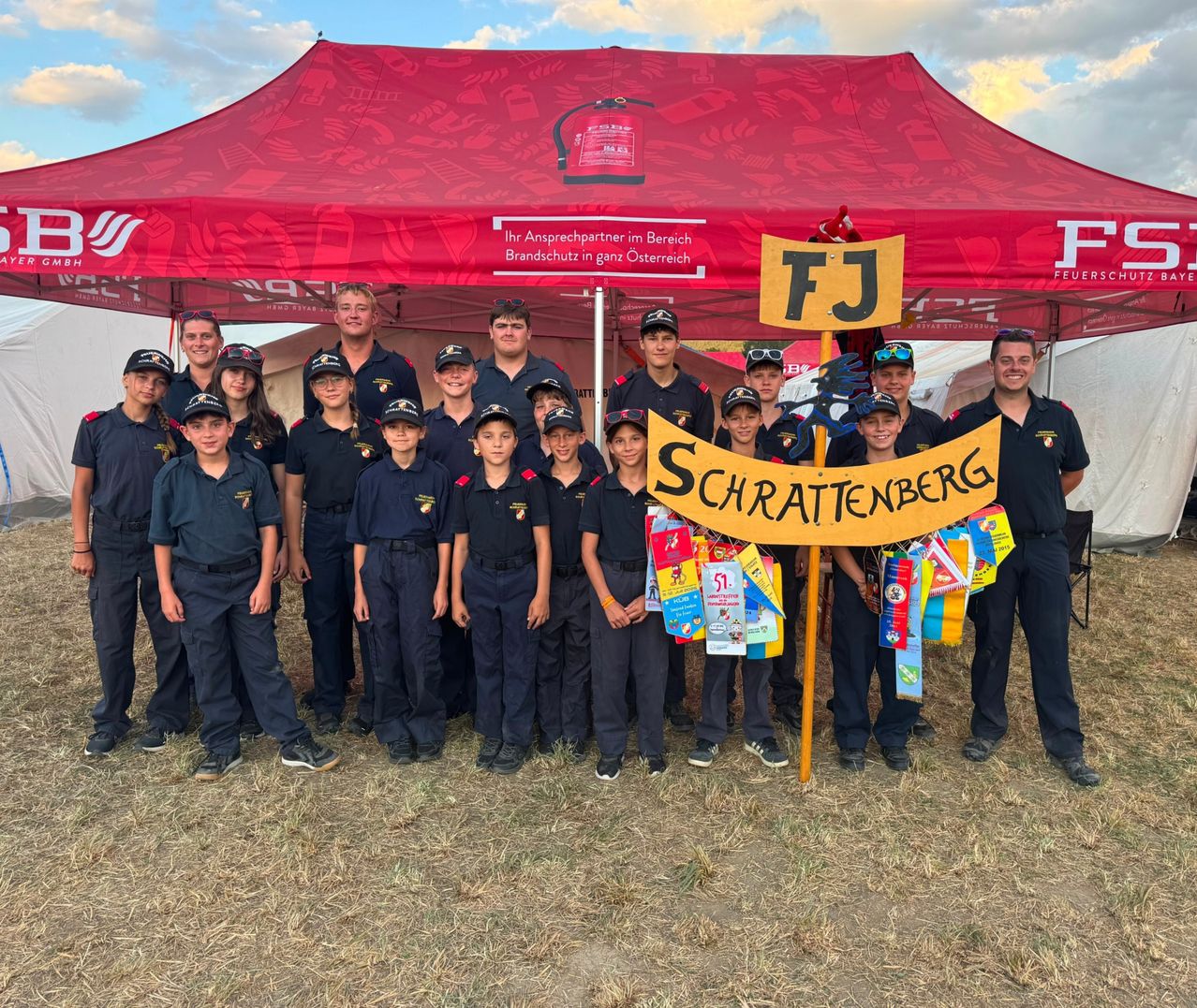 Eine Gruppe junger Menschen in Uniform, die unter einem roten Zelt mit einem Banner mit der Aufschrift Schrattenberg stehen. Sie halten ein gelbes Banner mit den Buchstaben FJ und der Nummer 51.
