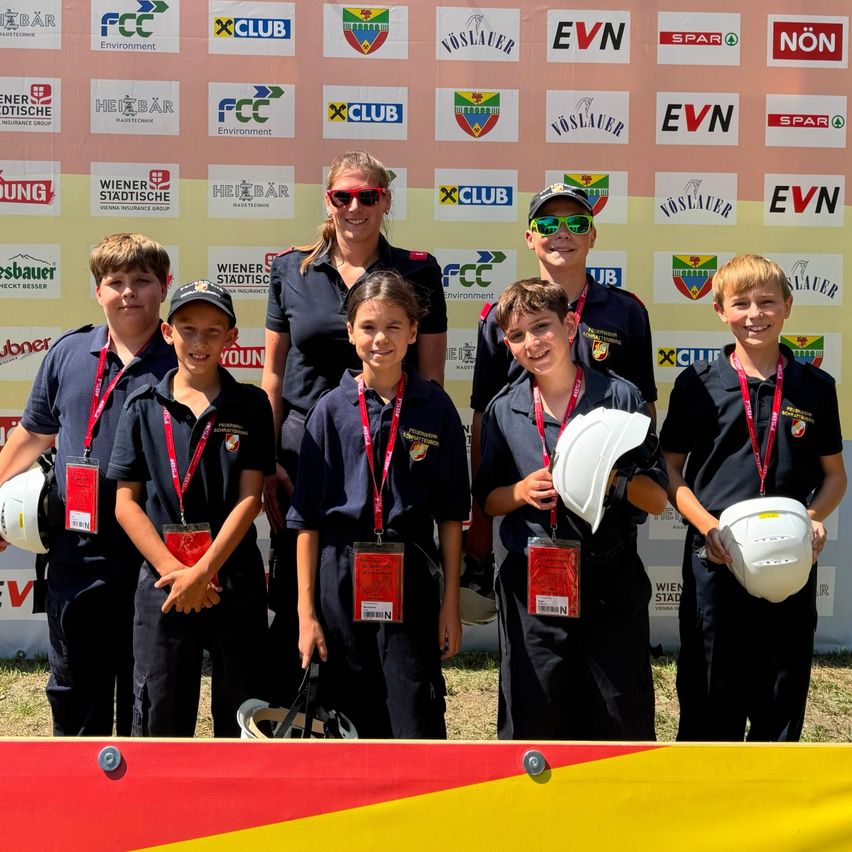 Eine Gruppe von Kindern und einer Frau in Uniformen und Helmen steht vor einer Wand mit verschiedenen Logos. Die Kinder lächeln und posieren für ein Foto.